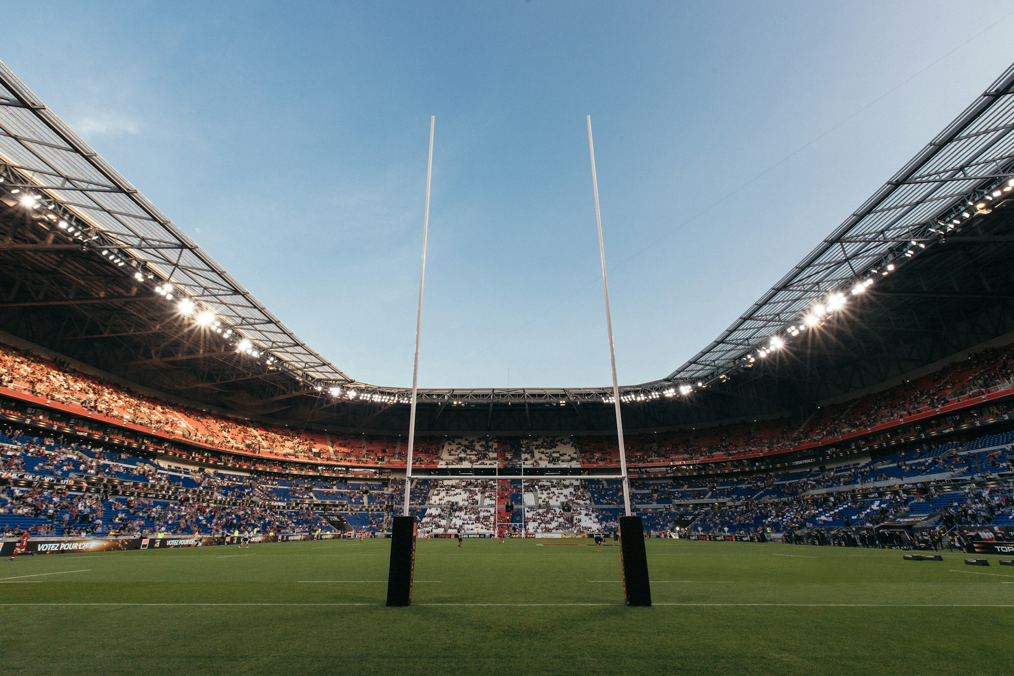 Stade de rugby vu depuis les poteaux d'en-but, tribunes garnies, projecteurs allumés, panneaux publicitaires en bord de terrain, joueurs au loin sous un ciel de fin de journée.